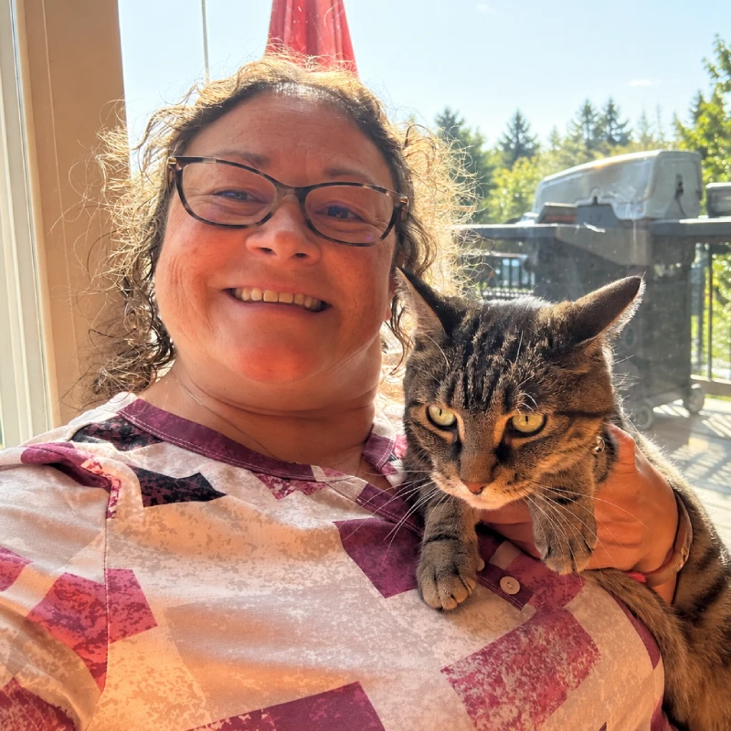 A smiling woman in glasses holds a tabby cat indoors beside a sunny window, with trees and a deck outside; the warm selfie feels cheerful and affectionate.